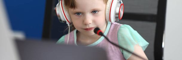 Маленькая девочка сидит за компьютером в наушниках с микрофоном
 - Фото, изображение