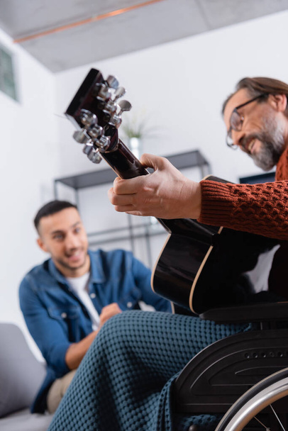 χαμηλή γωνία άποψη του ανάπηρου άνδρα που παίζει κιθάρα κοντά ισπανόφωνος γιος σε θολή φόντο - Φωτογραφία, εικόνα