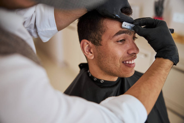 Счастливого улыбающегося мужчину в черном пеньюаре, который стрижется в парикмахерской - Фото, изображение