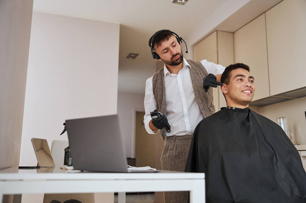 Cabeleireiro fazendo corte de cabelo com instrumento especial para cliente satisfeito em apartamentos - Foto, Imagem
