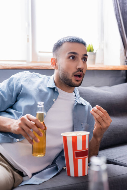 homme hispanique étonné avec bouteille de bière regarder la télévision et manger du pop-corn à la maison, avant-plan flou - Photo, image