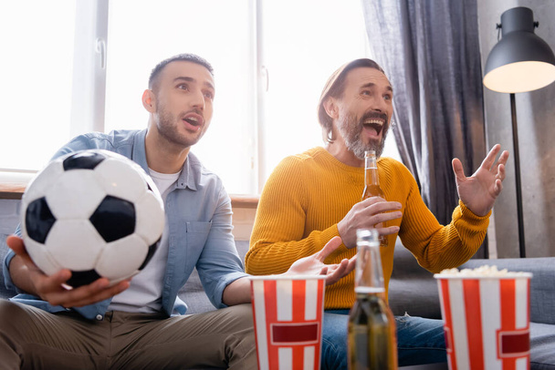 zdumiewający międzyrasowy ojciec i syn oglądający mistrzostwa piłki nożnej w pobliżu piwa i popcornu na niewyraźnym pierwszym planie - Zdjęcie, obraz