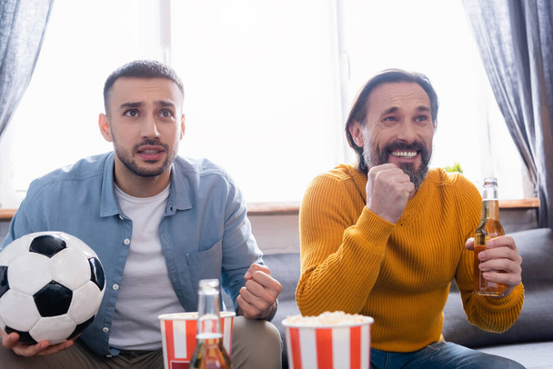 worried interracial father and son watching football match with beer and popcorn at home - Foto, Bild