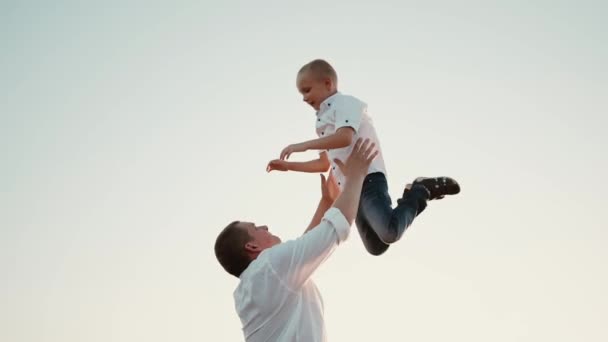 Apa feldob egy boldog fiút kék égen a levegőbe naplementekor. Apa és a kisgyerek játszanak, nevess. Családi utazások. A gyerek a szülő karjaiban mosolyog. Apa szabadnapja. A boldog család fogalma - Felvétel, videó