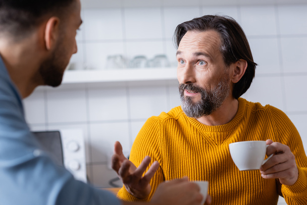 homem barbudo segurando xícara de café e gesticulando enquanto conversa com o filho na cozinha, foreground borrado - Foto, Imagem