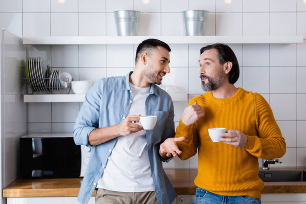 padre e hijo interracial sosteniendo tazas de café y haciendo gestos mientras hablan en la cocina - Foto, imagen