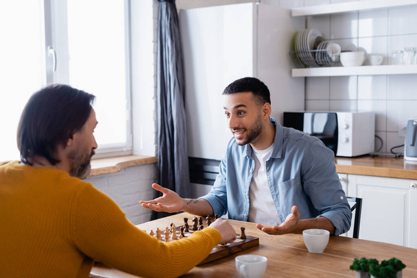 excited hispanic man gesturing while playing chess with father in kitchen - Photo, Image
