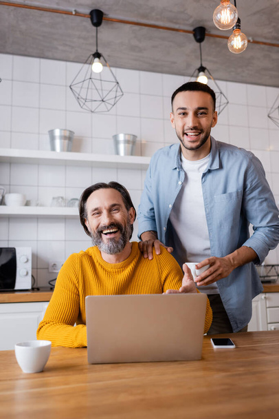 happy interracial father and son laughing at camera near laptop in kitchen - Photo, Image