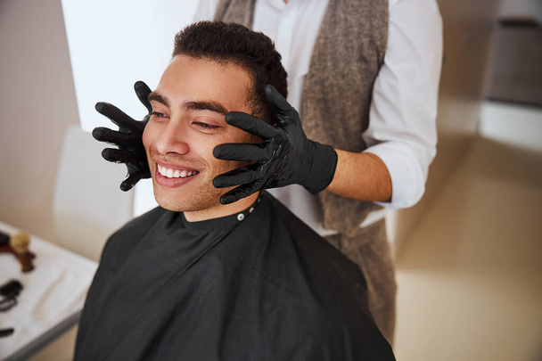 Κομμωτής βάζοντας χέρι στα αυτιά του πελάτη, ενώ ο επισκέπτης κοιτάζει μπροστά στο κουρείο - Φωτογραφία, εικόνα