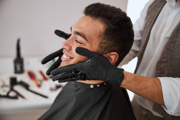 Handsome male resting in the barber shop while barber putting cosmetic oil on skin face - Photo, Image
