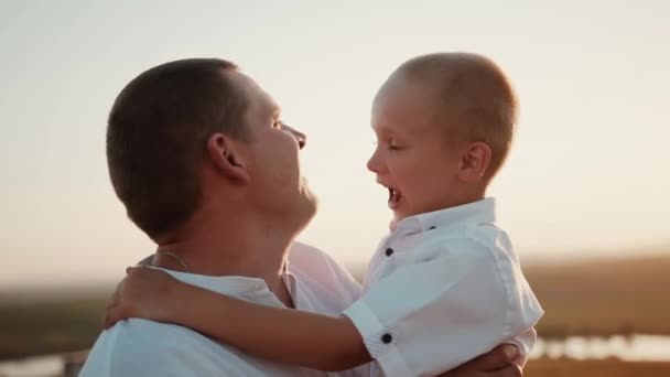Milující starostlivý otec při západu slunce drží v náručí veselého zábavného syna, který se pokouší kousnout otce do nosu, pohrávají si a šťastný muž objímá bezstarostné dítě a něžně ho políbí na tvář - Záběry, video