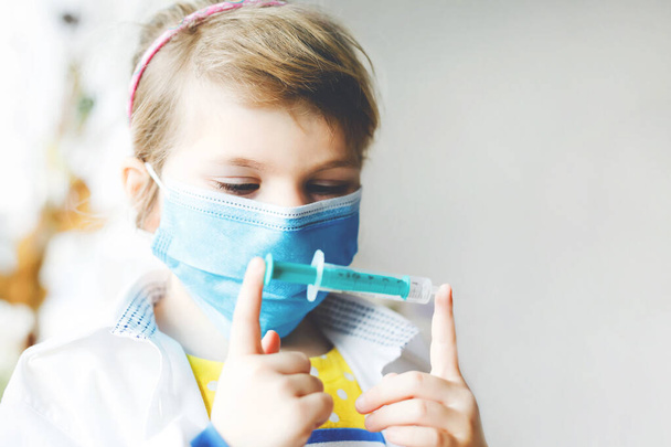 Mała dziewczynka robi zastrzyki. Słodkie dziecko z maską medyczną bawiące się w lekarza, trzymające strzykawkę ze szczepionką. Koncepcja szczepienia przeciw koronawirusowi. Kid grać rolę gry w domu. - Zdjęcie, obraz