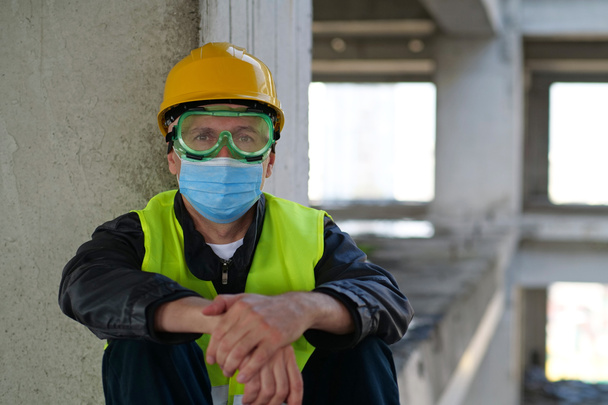 Travailleur en bleu masque de protection médicale, lunettes et casque jaune assis sur le chantier de construction et regarde la caméra - Photo, image