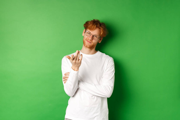 Jonge man met rood rommelig haar en bril, glimlachend blij en wijzend hand op camera, uw beurt gebaar, staande over groene achtergrond - Foto, afbeelding