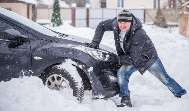 冬、人々と車の問題の概念。男は車を押すと、雪の中で立ち往生しようとします。相互扶助。冬の問題だ。輸送、冬と車のコンセプト-雪の中に閉じ込められた車を押す男 - 写真・画像