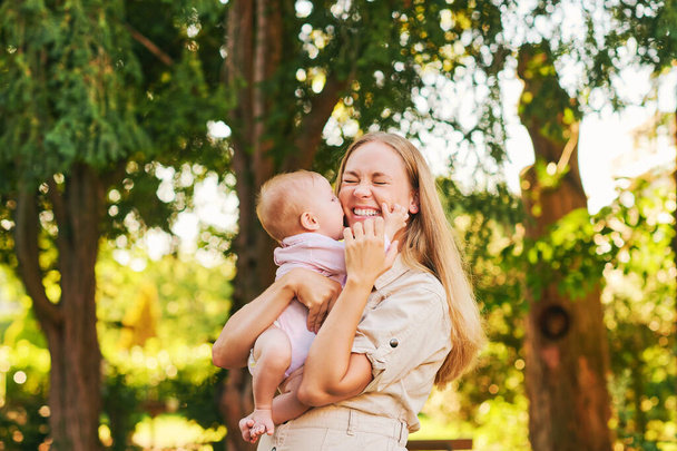 Αξιολάτρευτο μωρό να φιλάει τη μαμά της, νεαρή χαρούμενη γελαστή μητέρα στο καλοκαιρινό πάρκο  - Φωτογραφία, εικόνα