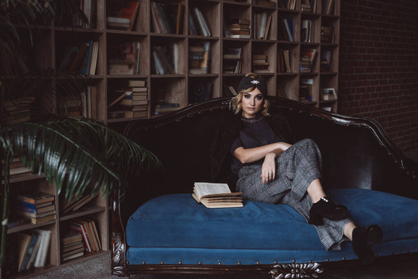 Beautiful stylish blonde woman in a knitted headband, jacket and culottes in a loft interior with a shelf with books. Soft selective focus, defocus. No retouching. - Photo, Image