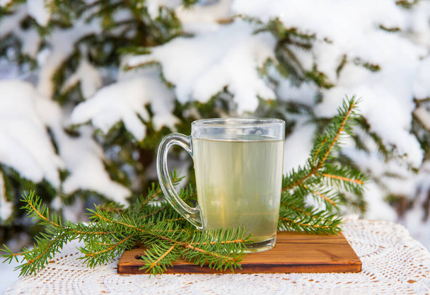 Spruce tree needle tea infusion in transparent glass tea cup. Snowy blurred spruce tree on background, outdoors on cold winter day. Room for text. - Photo, Image