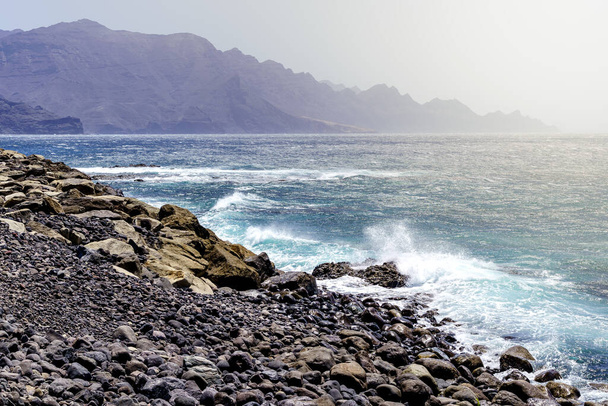 Paisaje marino con playa rocosa y olas rompiendo en la costa, montañas distantes con altos acantilados. - Foto, Imagen