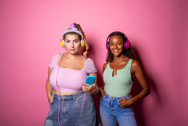 Fit und überdimensioniert posiert junge Erwachsene auf rosa Hintergrund und hört Musik - Diverse Frauen mit unterschiedlichen Körperformen isoliert auf Hintergrund - Selbstwertgefühl, Selbstakzeptanz, körperpositives Konzept - Foto, Bild