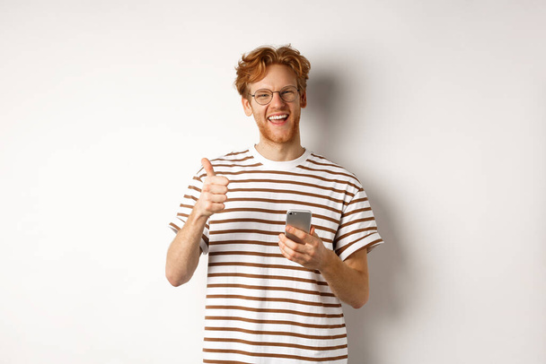 Технология и концепция электронной коммерции. Удовлетворенный мужчина модель с рыжими волосами, показывая большие пальцы вверх и держа смартфон, белый фон - Фото, изображение