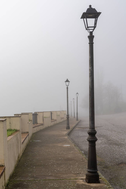 Ένας αστικός δρόμος την ημέρα με πολύ πυκνή ομίχλη και ομίχλη και μια σειρά από νοσταλγικές παλιές μαύρες λάμπες του δρόμου - Φωτογραφία, εικόνα