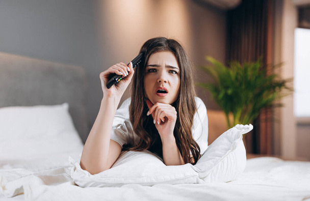 Женщина в белой шелковой пижаме смотрит телевизор в спальне на кровати. Женщина-модель с пультом от телевизора в руках лежит на кровати. утром или вечером в спальне с моделью - Фото, изображение