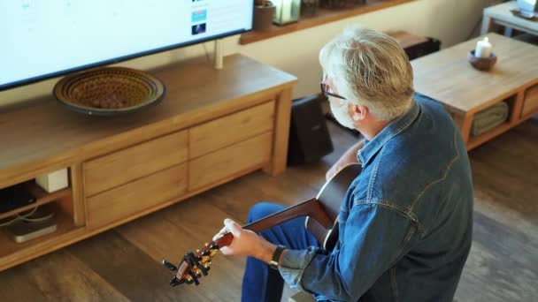 Vista posterior de hombre caucásico senior relojes lección en línea en el monitor grande y toca la guitarra. Distanciamiento social en cuarentena. Imágenes de alta calidad 4k - Imágenes, Vídeo