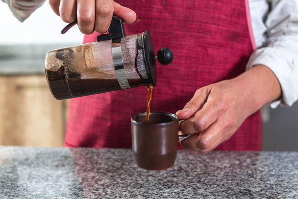 Großaufnahme der Hände von jemandem in roter Schürze, der eine in einer französischen Presse gebrühte Tasse Kaffee einschenkt. - Foto, Bild