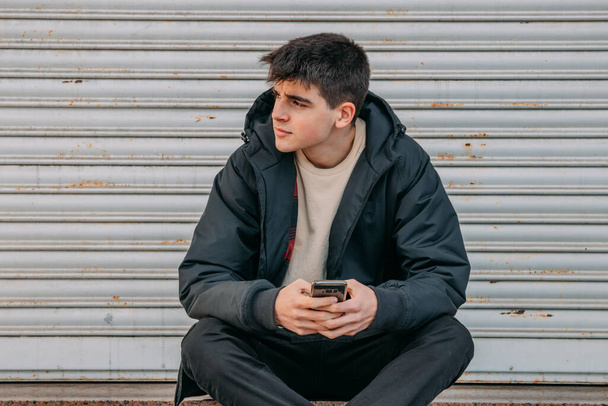 νεαρός άνδρας με κινητό τηλέφωνο κάθεται στο δρόμο της πόλης - Φωτογραφία, εικόνα
