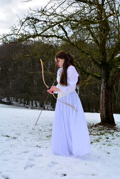 Mujer joven de pie de lado con vestido blanco largo en la nieve frente a un bosque, jugueteando con un arco y flecha - Foto, Imagen