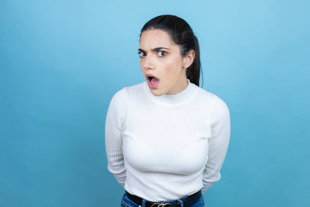 Mujer joven caucásica vistiendo suéter blanco sobre fondo azul asustada y conmocionada con expresión sorpresa y asombrada, miedo y cara excitada. - Foto, imagen