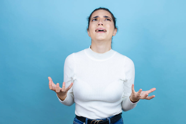 Young caucasian woman wearing white sweater over blue background crazy and mad shouting and yelling with aggressive expression and arms raised. Frustration concept. - Photo, Image