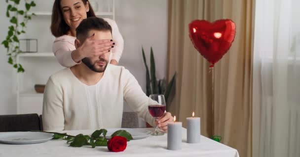Zbliżenie mężczyzna czeka przy stole ze świecami w restauracji w domu czekając jego ukochanej żony dotykając szklanki czerwonego wina, kobieta zamykając oczy do męża i dając czerwone punkty prezent palec na złotej wstążce - Materiał filmowy, wideo