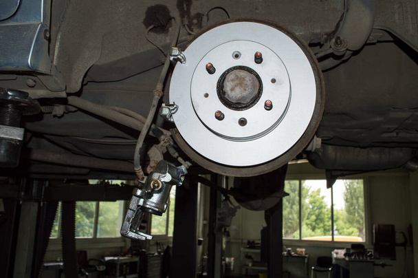 Vista lateral del nuevo disco de freno y pinza de rueda trasera montados en un coche gris que cuelga en un ascensor en el taller de reparación de automóviles - Foto, imagen