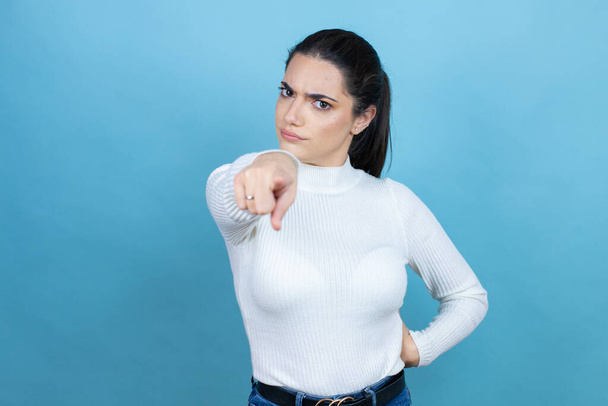 Joven mujer caucásica vistiendo suéter blanco sobre fondo azul señalando con el dedo a la cámara y a ti, gesto confiado mirando serio - Foto, imagen