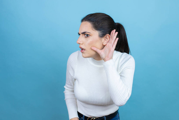 Jeune femme caucasienne portant un pull blanc sur fond bleu surpris avec la main sur l'oreille en écoutant une rumeur ou des commérages - Photo, image