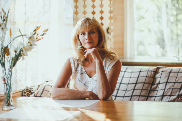 Portrét elegantní atraktivní blondýny bělošky, stařeny se stylovými vlasy v bílém tričku, sedící u jídelního stolu u okna a dívající se na kameru s veselým úsměvem. slunečné počasí. - Fotografie, Obrázek