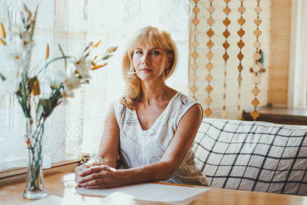 Portrait der eleganten attraktiven blonden Kaukasierin, einer älteren Frau mit stylischem Haar in weißem T-Shirt, die an einem Esstisch am Fenster sitzt und mit fröhlichem Lächeln in die Kamera blickt. sonniges Wetter. - Foto, Bild