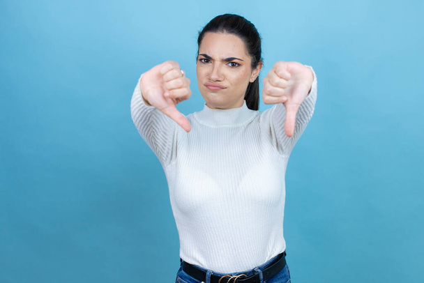 Jeune femme caucasienne portant un pull blanc sur fond bleu avec un visage en colère, signe négatif montrant aversion avec les pouces vers le bas - Photo, image