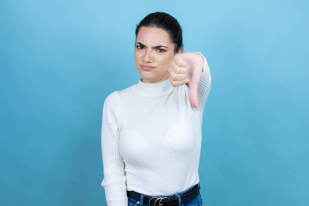 Young caucasian woman wearing white sweater over blue background with angry face, negative sign showing dislike with thumb down - Photo, Image