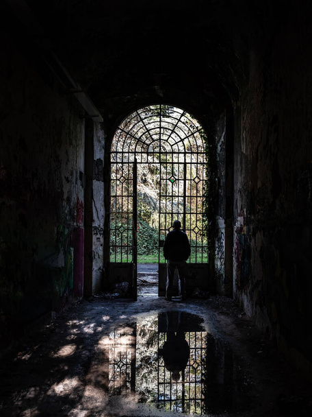 Silhouette of a man observing the outside world looking from the grate of a door and his image is reflected in a puddle inside an abandoned building - Photo, Image