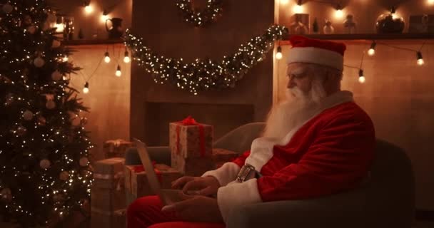 Santa Claus sits next to a Christmas tree on a comfortable soft sofa, holds a laptop in his lap and answers emails from young children. - Footage, Video