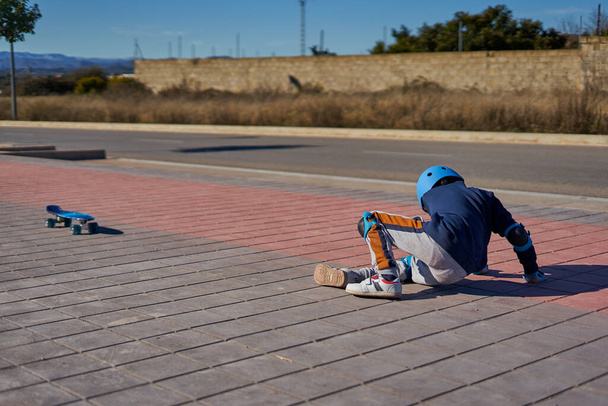 chico levantándose después de caer en un accidente de skate. Lleva casco y protecciones y está tirado en un carril bici. - Foto, Imagen