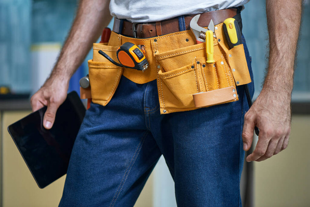Cropped shot of young repairman wearing a tool belt with various tools holding digital tablet while standing indoors - Photo, Image