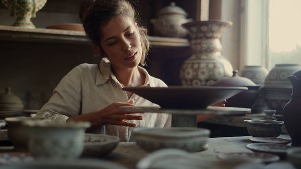 Señora sonriente tocando plato en cerámica. Artista disfrutando de producto de arcilla en taller - Foto, Imagen