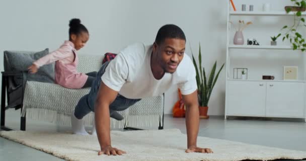 Afro-American single-parent family having fun at home, strong father doing sports during lockdown pushing up from floor on carpet while unexpectedly beloved daughter jumps on back of her daddy hugs - Footage, Video