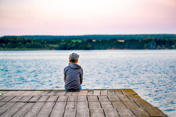 Boy sits on wooden bridge near a pond. Rear view from a back on small boy. Beautiful landscape. - Φωτογραφία, εικόνα