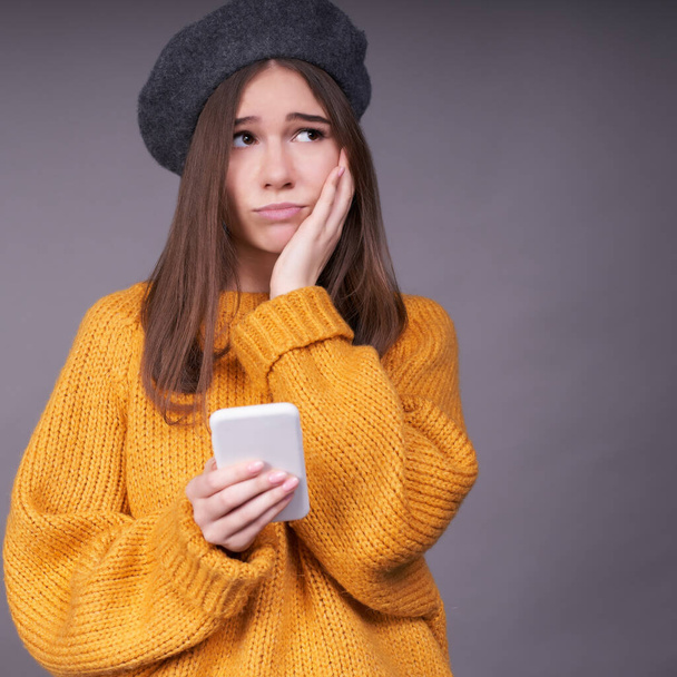 Vertical shot, a sad young Caucasian girl with fair hair, in a yellow sweater and a gray beret, holds a smartphone in her hands, looks away, holds her hand on her cheek, learned the sad news, is sad. - Fotoğraf, Görsel