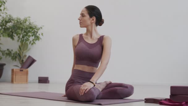 Weite Aufnahme einer schlanken jungen kaukasischen Frau in Sportbekleidung, die in Lotusposition auf einer Yogamatte im Haus sitzt und ihren Rücken aufwärmt - Filmmaterial, Video
